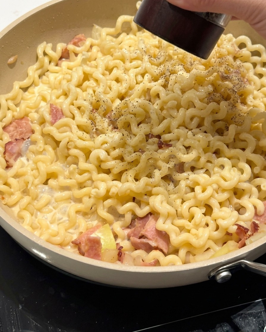 Eine Zange mischt die cremigen Spiralnudeln in einer Pfanne mit Speck und Soße.