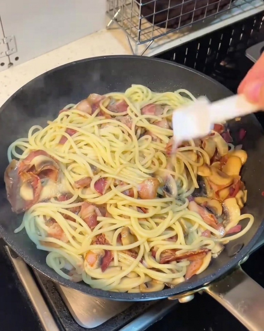 Ein Hand hält einen Pfefferstreuer über die Pfanne mit Spaghetti und Speck.
