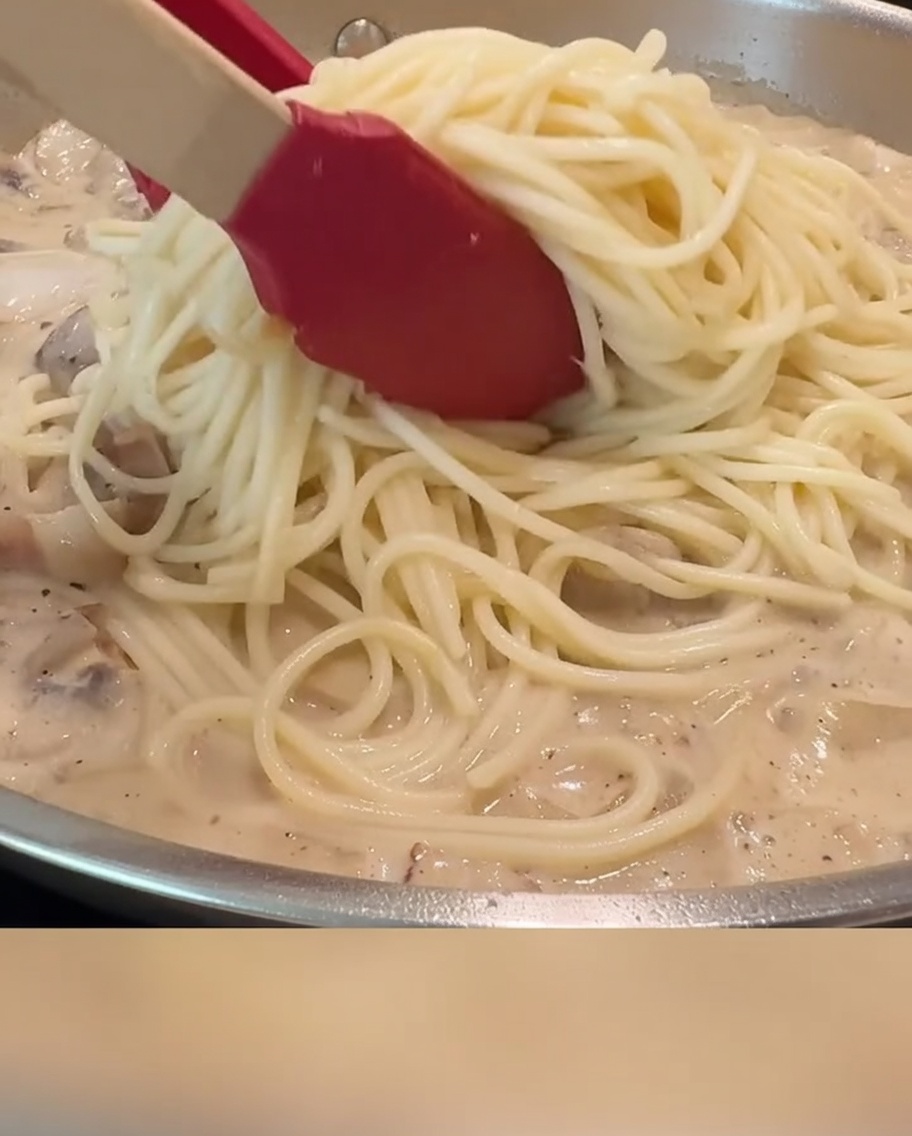 Spaghetti werden mit roter Zange mit cremiger Soße, Speck und Champignons in einer Pfanne vermengt.