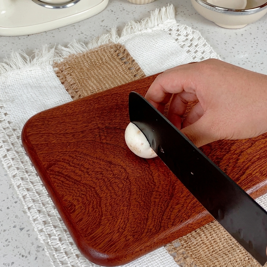 Eine Hand schneidet mit einem schwarzen Messer einen weißen Champignon auf einem Holzbrett.