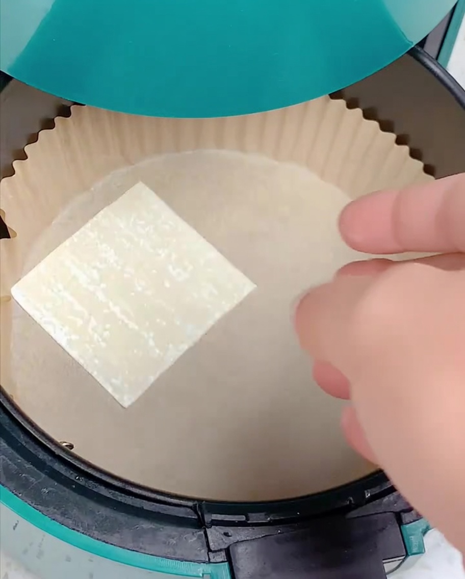 Eine Hand legt ein quadratisches Wonton-Teigblatt in einen mit Backpapier ausgelegten Air-Fryer-Korb.