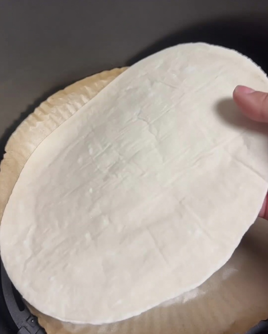 Ein roher Frühlingszwiebel-Pfannkuchen liegt flach in einem mit Backpapier ausgelegten Airfryer-Korb.