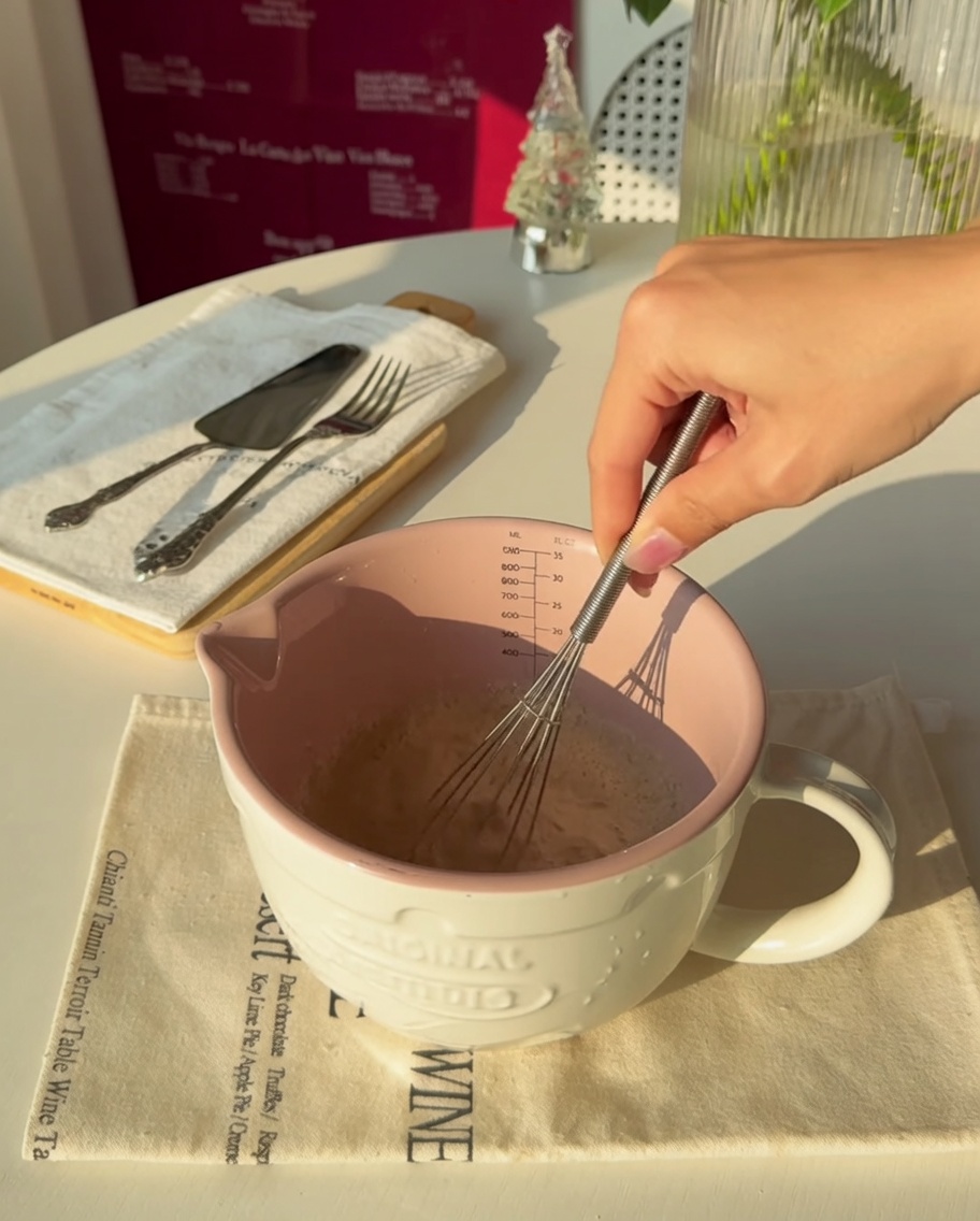 Nahaufnahme einer Hand, die Hefe in warmes Wasser in einem rosa Messbecher rührt.
