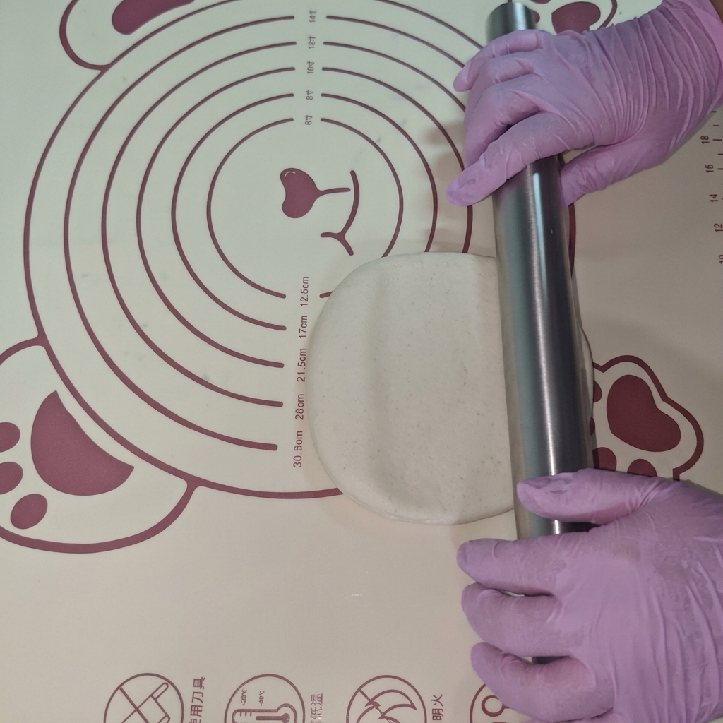 Ein Stück Teig wird auf einer Silikonmatte mit einem Nudelholz zu einem flachen Kreis ausgerollt.