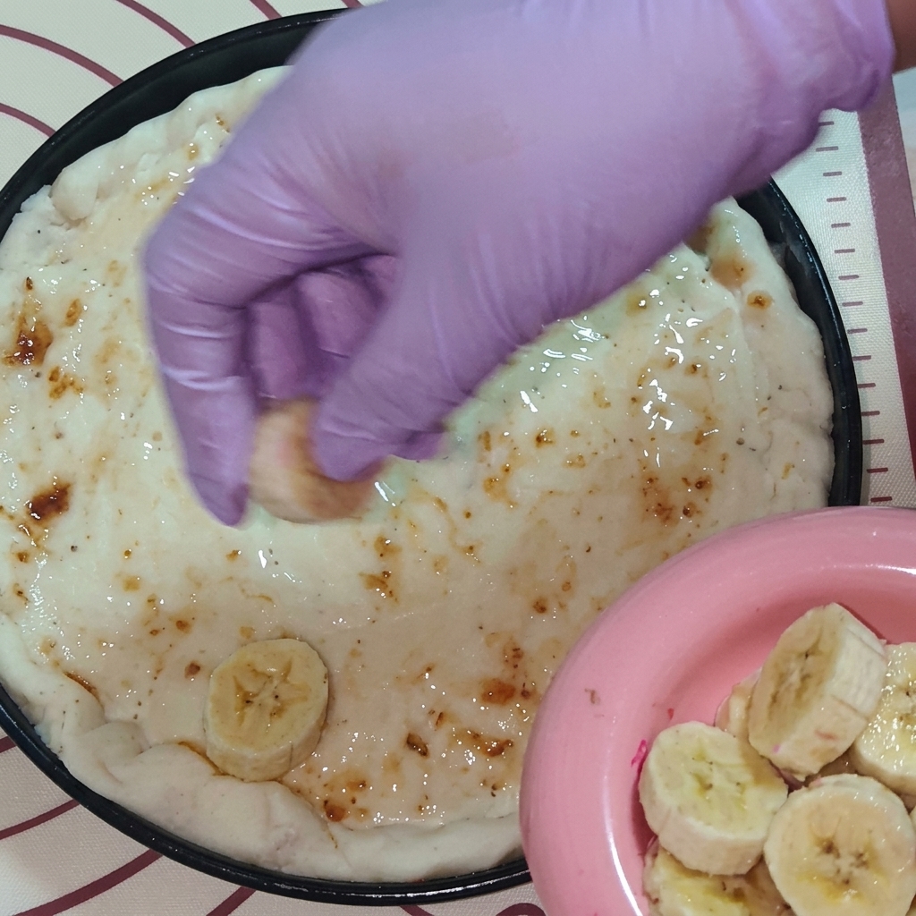 Eine Hand in einem lila Handschuh legt frische Fruchtscheiben, darunter Banane und Drachenfrucht, auf den mit Marmelade bestrichenen Pizzateig.