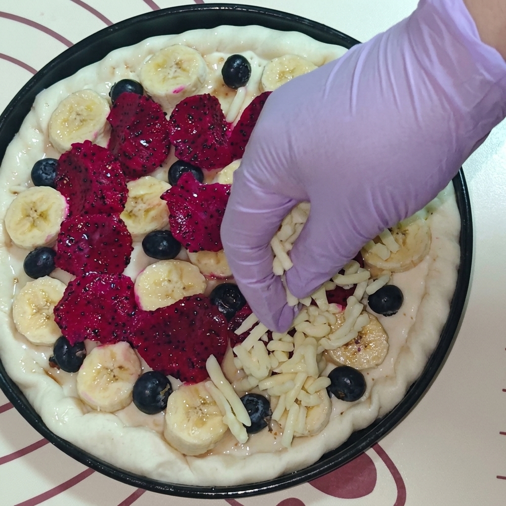 Draufsicht auf eine Hand in einem lila Handschuh, die geriebenen Mozzarella über die Fruchtpizza streut.