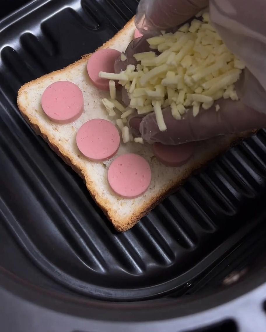 Geriebener gelber Käse wird über die Würstchenscheiben auf dem Toast gestreut.