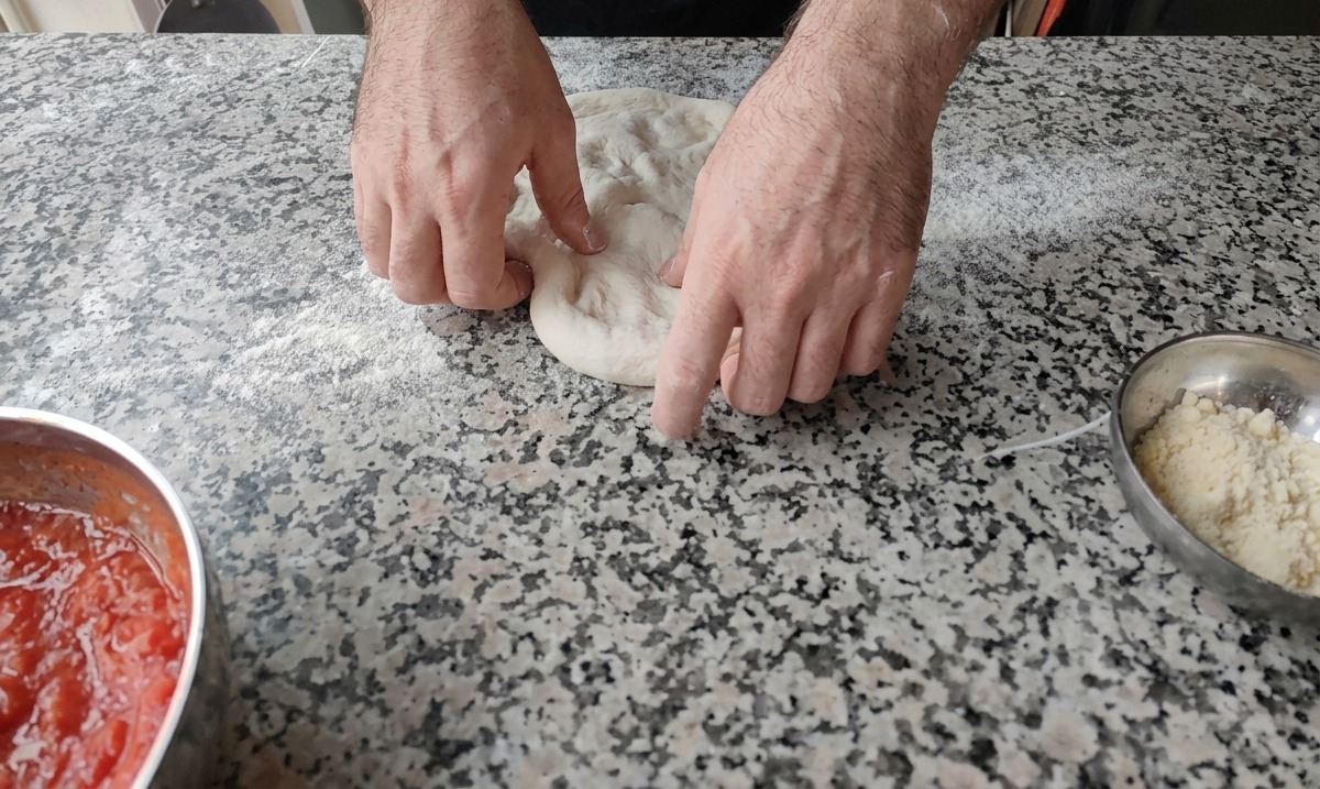 Zwei Hände drücken den Teigball auf einer bemehlten Arbeitsfläche flach.