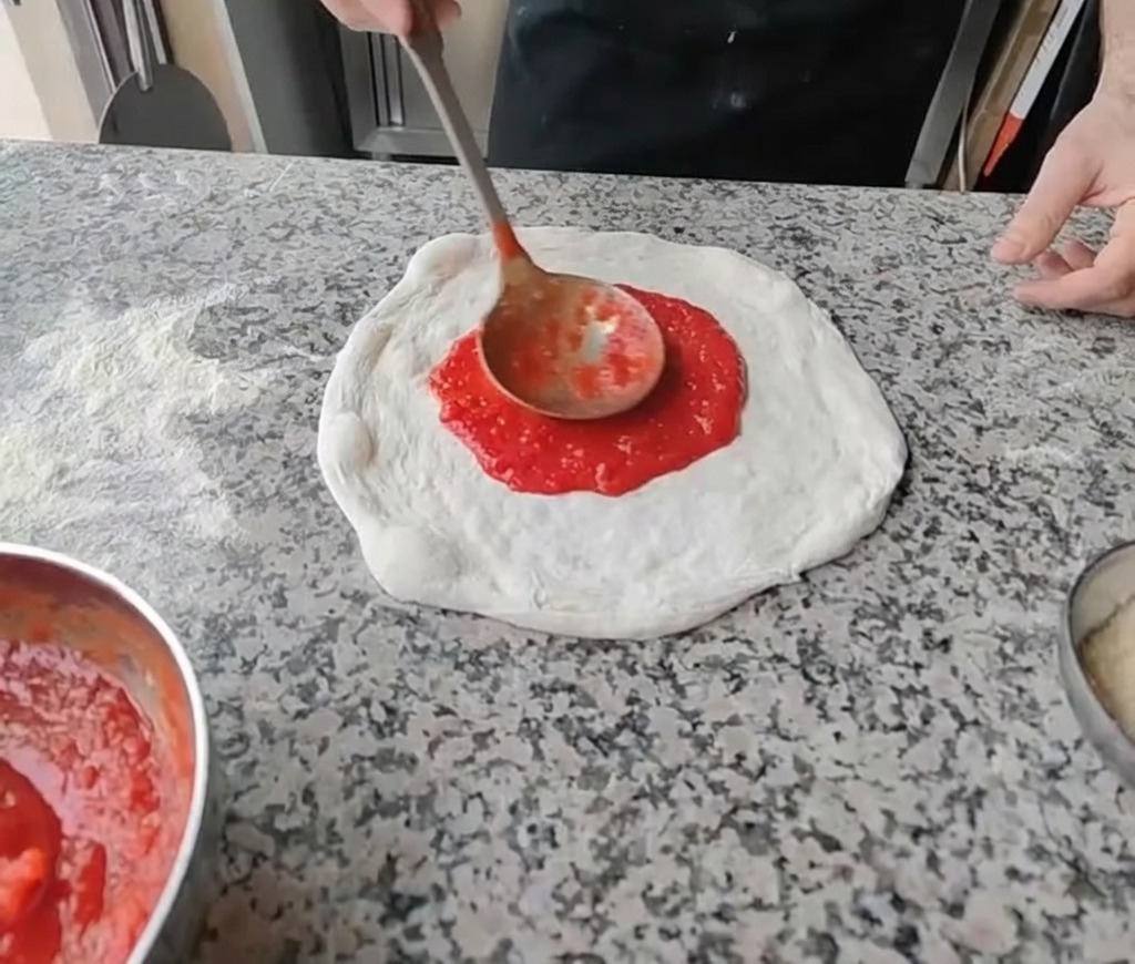Die Tomatensoße wird mit einer Schöpfkelle in kreisenden Bewegungen auf den Teig gegeben.
