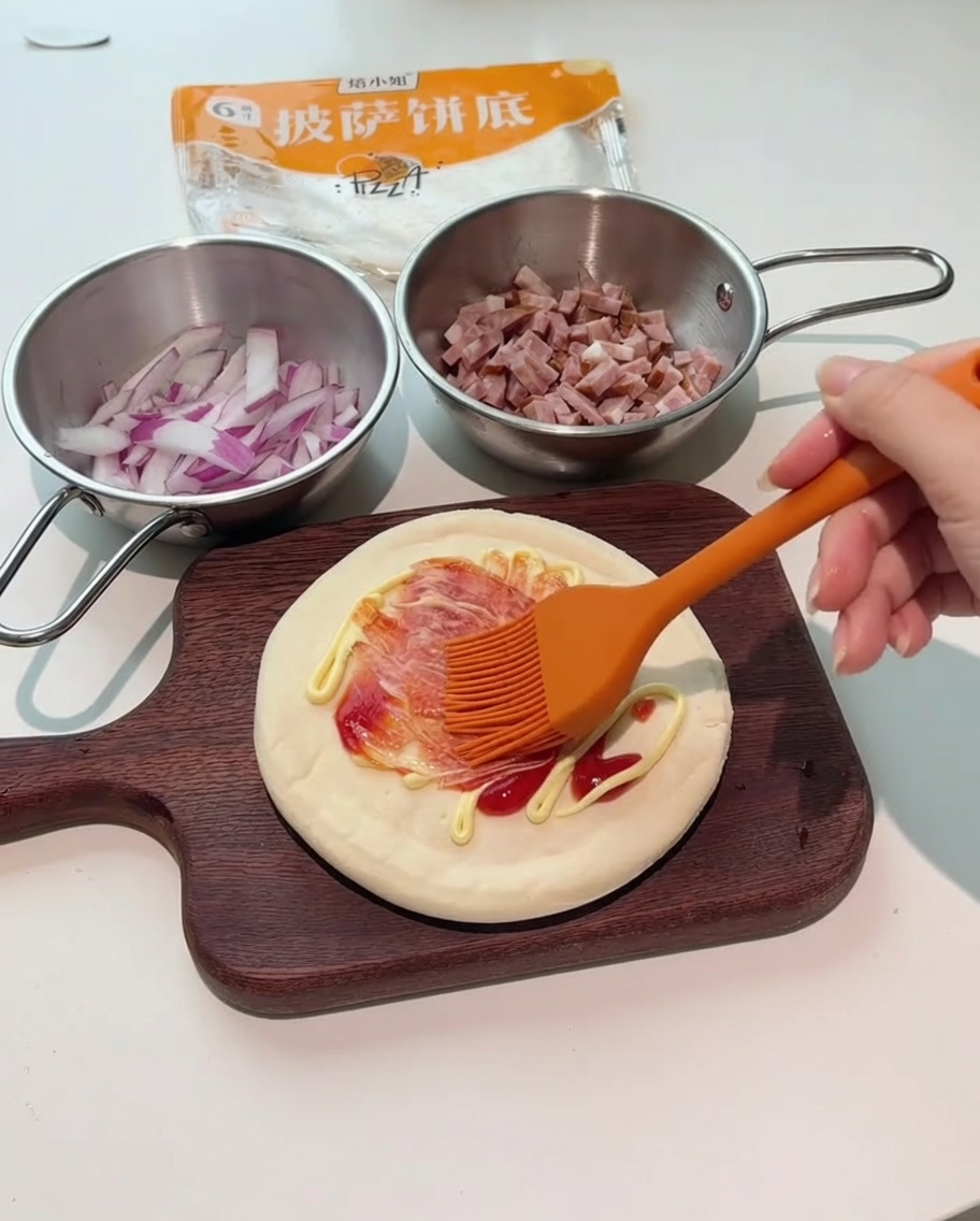 Ein orangefarbener Silikonpinsel verteilt rote und weiße Saucen auf dem Pizzaboden.