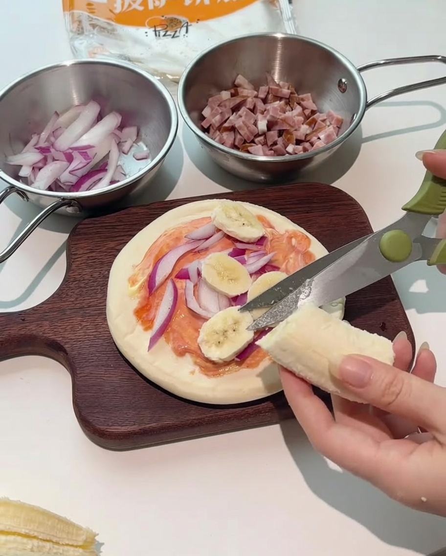 Eine Küchenschere schneidet frische Bananenscheiben direkt auf den Pizzabelag.