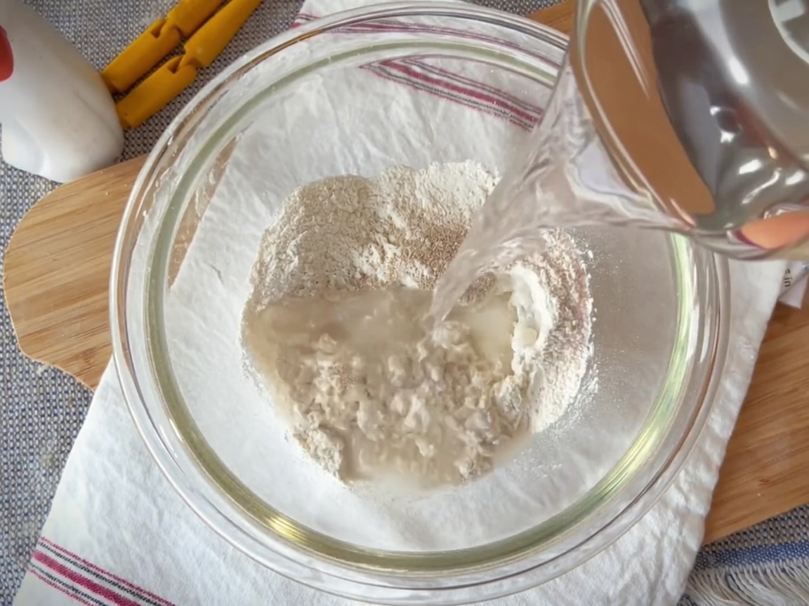 Eine Glasschüssel mit einer Mischung aus Mehl, Hefe, Salz und Wasser, die mit einem Spatel vermengt wird.