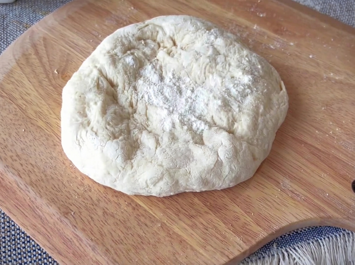 Ein rundes Stück Pizzateig wird auf einer Holzoberfläche geformt, mit einem deutlich erkennbaren dicken Rand.