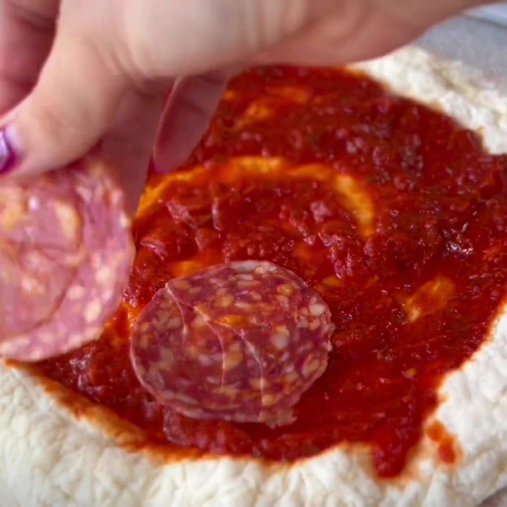Salamischeiben werden vorsichtig auf den bestrichenen Bereich des Pizzateigs gelegt.