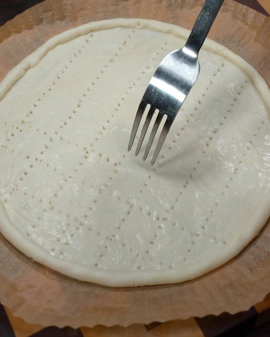 Eine Metallgabel drückt in den rohen Teigboden, um ein Gitter aus kleinen Einstichlöchern zu erzeugen.