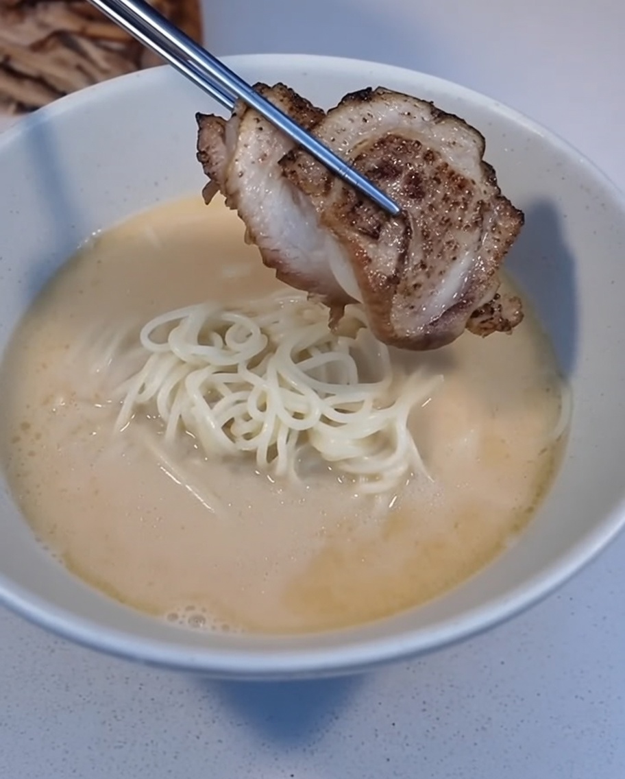 Eine Scheibe angebratenes Char Siu wird mit einer Pinzette auf eine Schüssel Ramen gelegt.