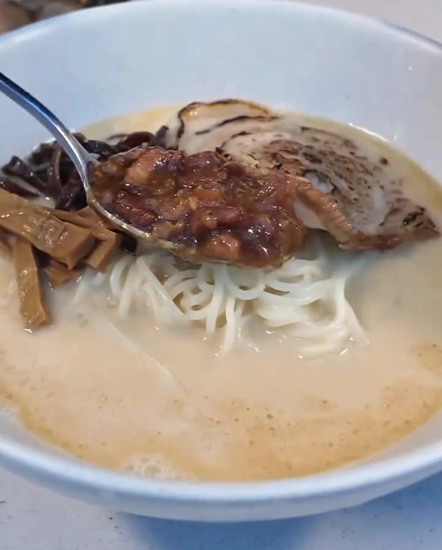 Ein Löffel geschmorte Fleischsauce wird neben der weichgekochten Eihälfte in die Ramen-Schüssel gegeben.