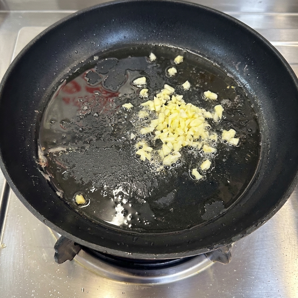 Fein gehackter Knoblauch, der leicht sprudelnd in einer dünnen Schicht heißem Öl in einer schwarzen Pfanne brät.