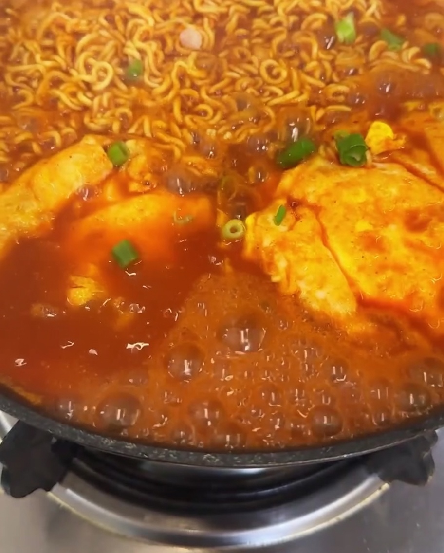 Nahaufnahme eines Topfes mit Kimchi-Ramen, garniert mit frisch gestreuten Frühlingszwiebeln und leuchtend roten Chilischeiben, während die Brühe köchelt.