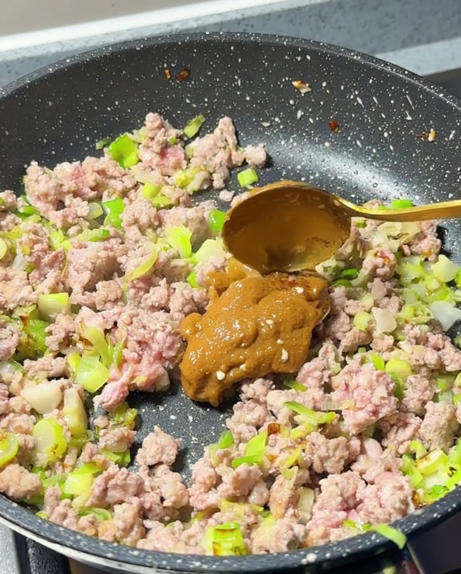Ein Löffel hellbraune Miso-Paste wird zum gebratenen Hackfleisch in die Pfanne gegeben.
