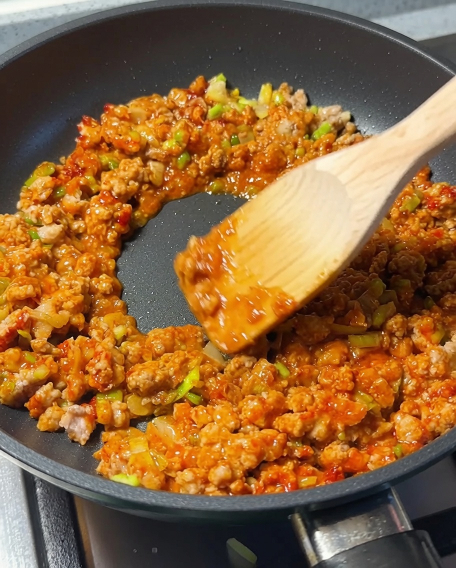 Ein Holzlöffel rührt das Hackfleisch um, während Sojasauce in die Pfanne gegeben wird.