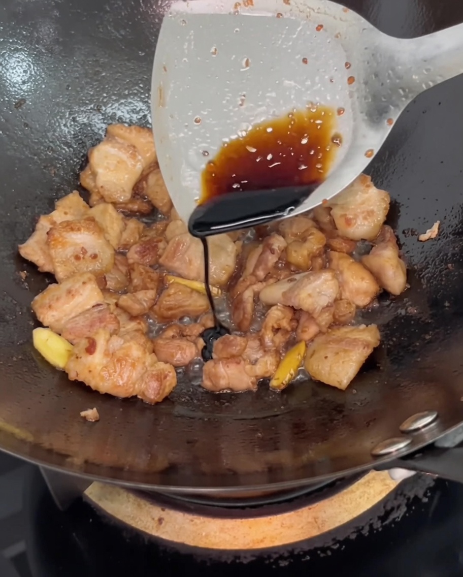 Dunkle Sojasauce wird von einem Pfannenwender über den schmorenden Schweinebauch geträufelt.