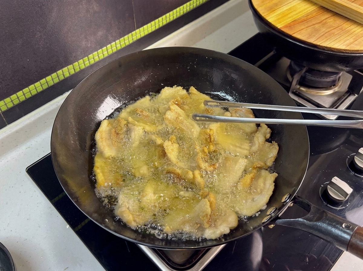 Mehrere Schweinebauchscheiben frittieren im Wok und werden mit einer Zange gewendet.