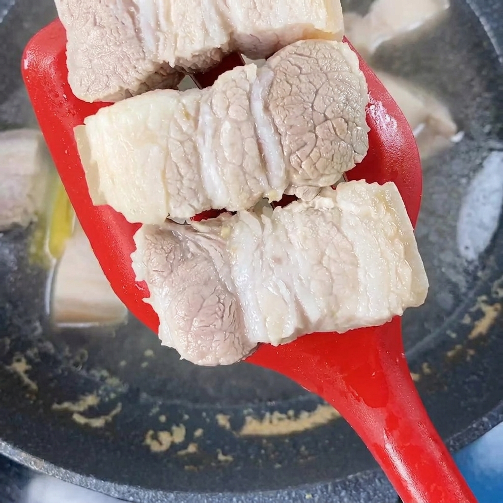 Blanchierte Schweinebauchstücke werden mit einem roten Pfannenwender aus kochendem Wasser gehoben.