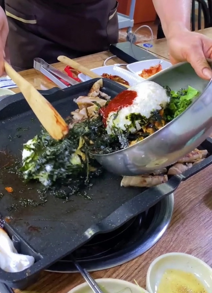 Reis, Gemüse und Algen werden aus einer Metallschüssel auf einen Grill mit Schweinefleisch gegeben.