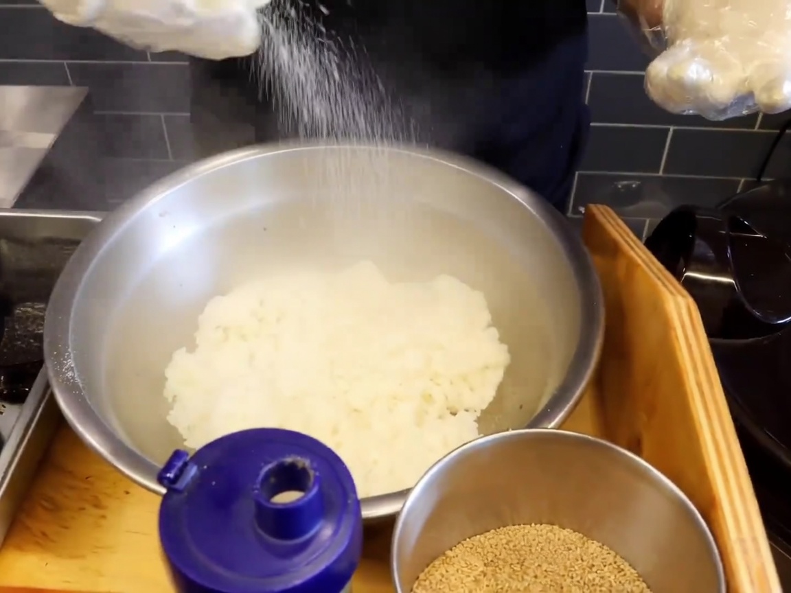 Nahaufnahme einer großen Edelstahlschüssel mit dampfendem Klebreis, bestreut mit Salz und Sesam.