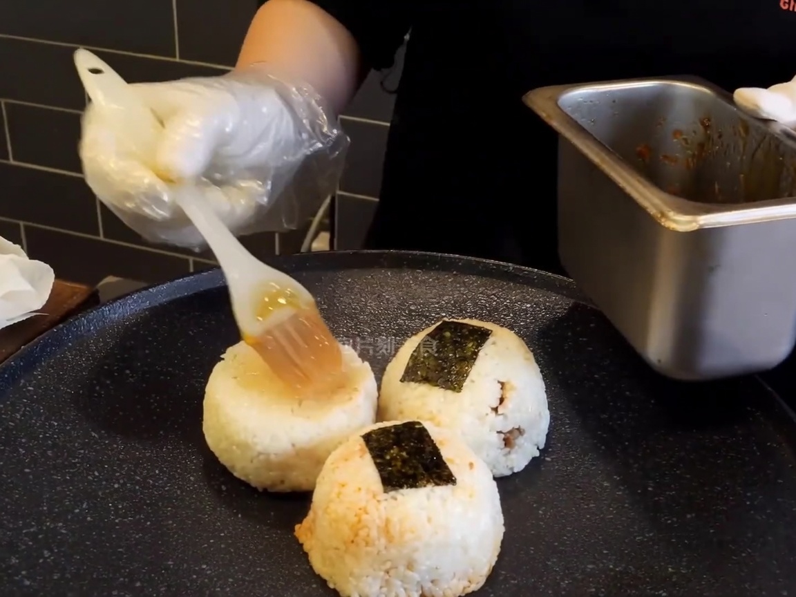 Reisbällchen auf einer heißen Grillplatte werden mit einer braunen Soße bepinselt.