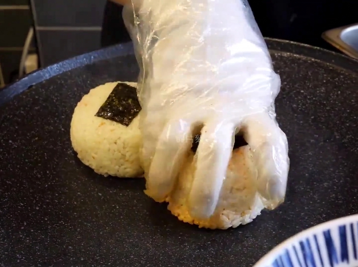 Eine behandschuhte Hand drückt die Reisbällchen auf einer schwarzen Grillplatte an.