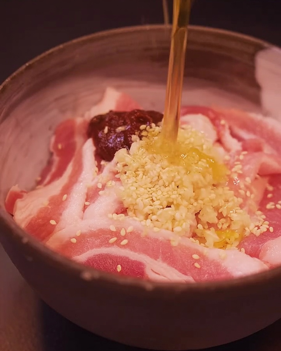 Schweinebauchscheiben in einer Schüssel, die mit Gochujang, Knoblauch, Sesamsamen und Sesamöl gewürzt werden.