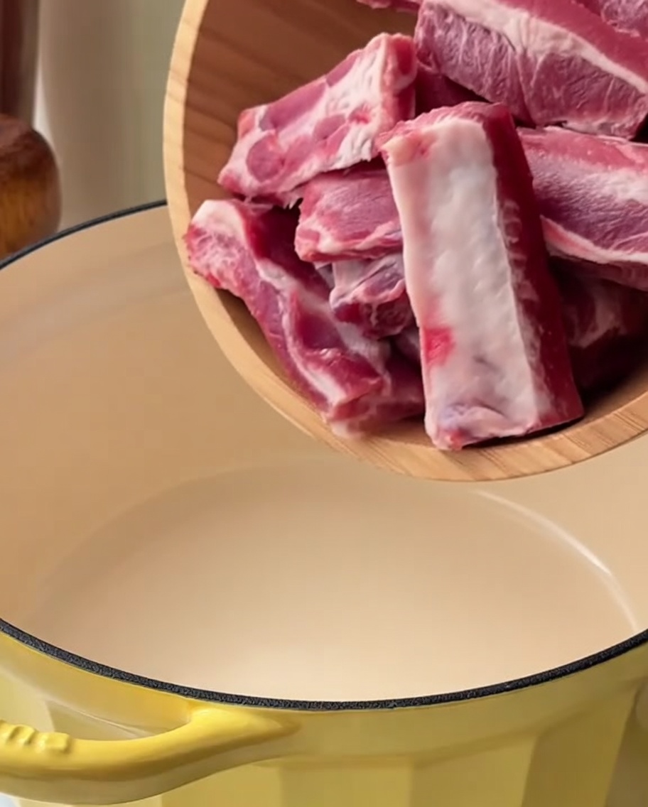 Schweinerippchenstücke liegen in einem gelben Topf in Wasser mit Frühlingszwiebeln und Ingwer.