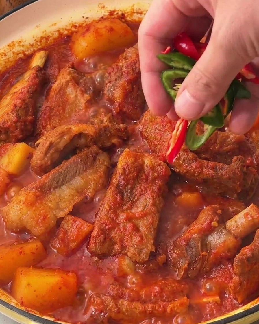 Eine Hand streut frische grüne und rote Chilistücke über den fertigen koreanischen Schweinerippchen-Eintopf.