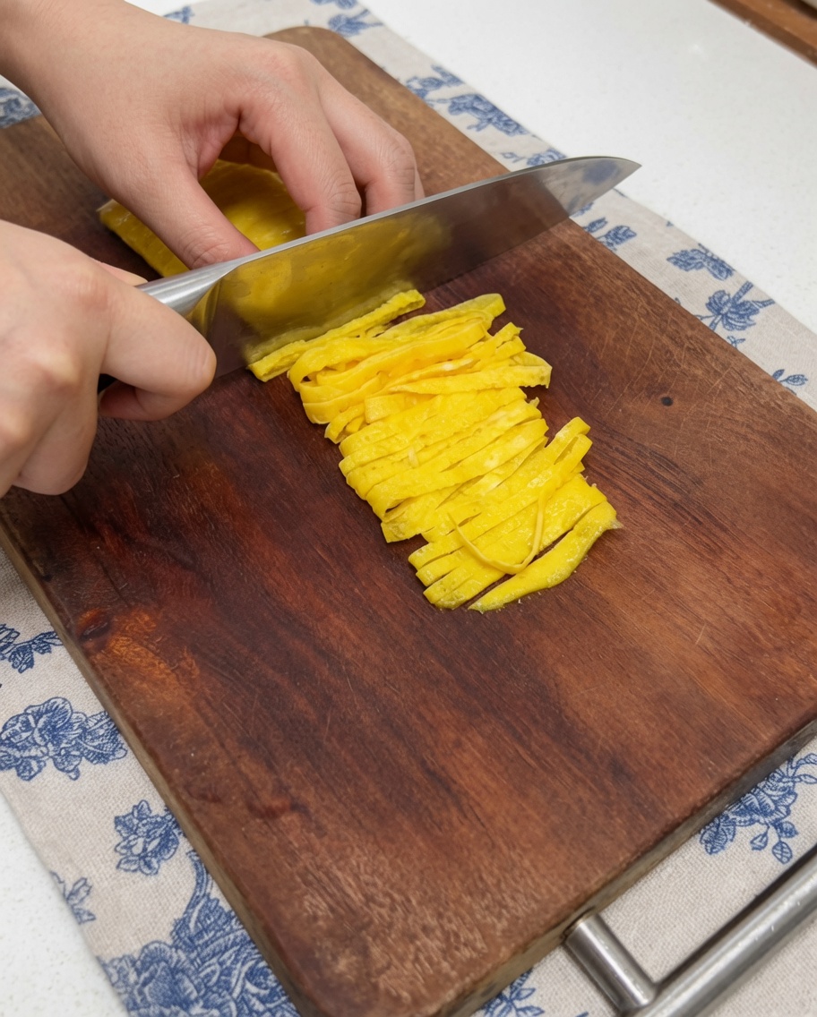 Nahaufnahme einer Hand, die mit einem Küchenmesser ein dünnes Eier-Crêpe in Streifen schneidet.