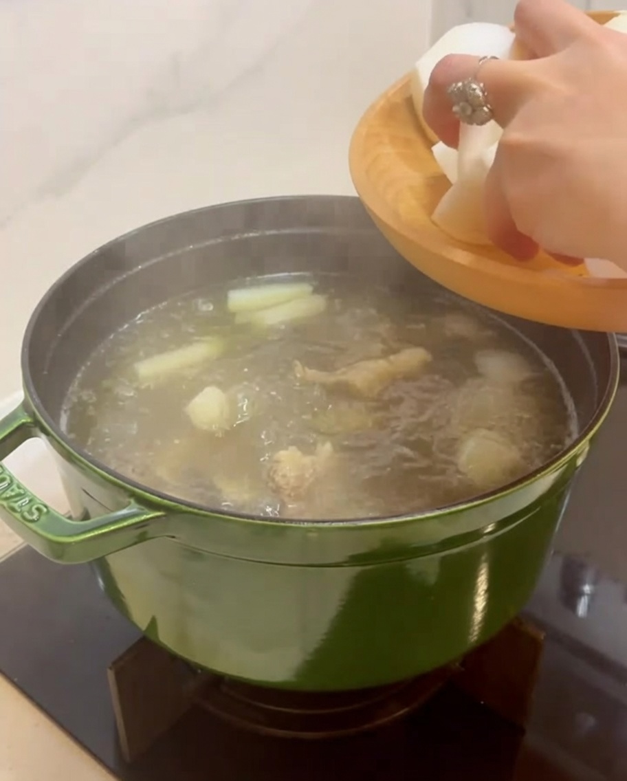 Hand gibt große, weiße Stücke von geschältem koreanischem Rettich in einen köchelnden Topf mit Rindersuppe.