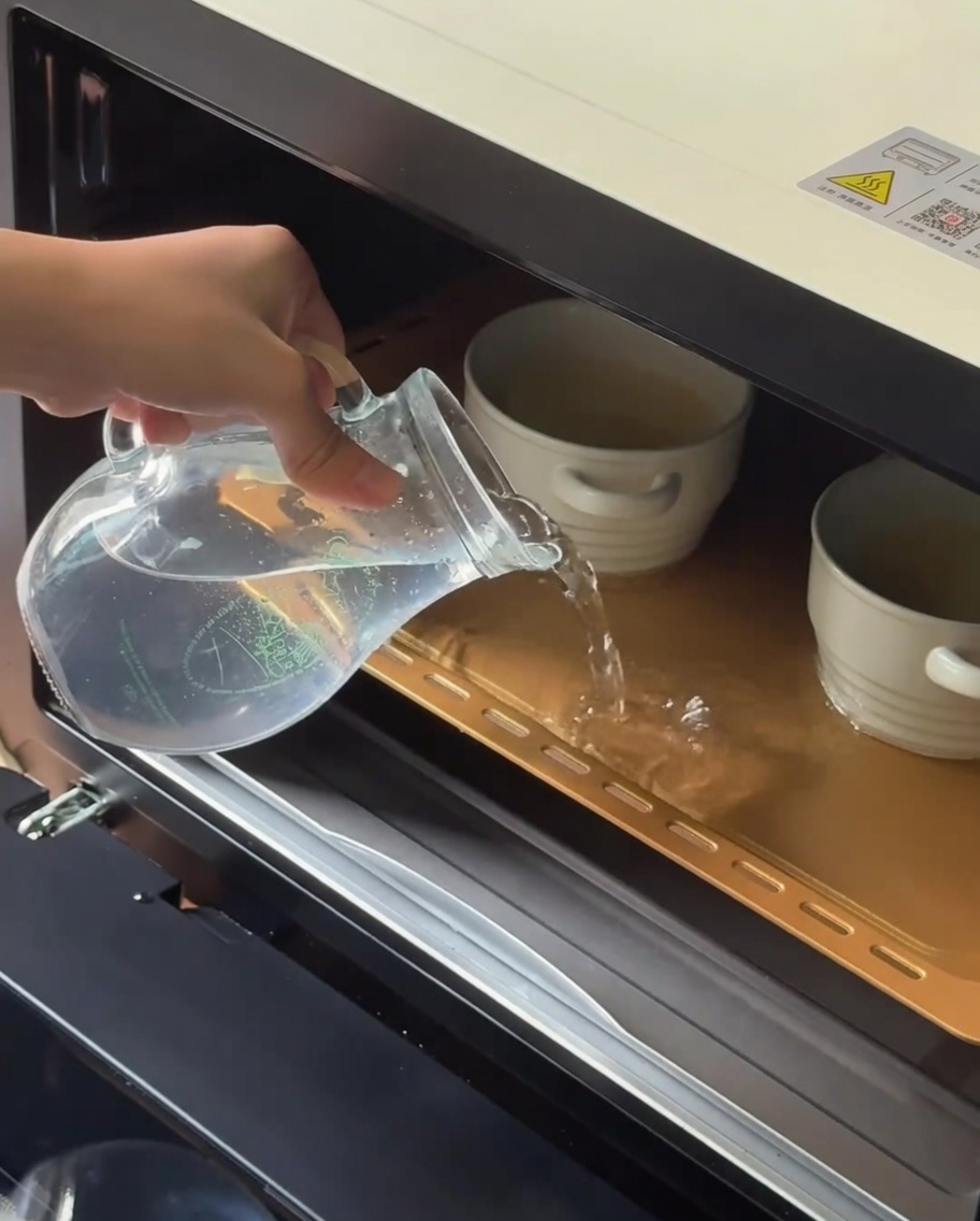 Warmes Wasser wird aus einem Glaskrug in ein Backblech mit Keramik-Förmchen gegossen.