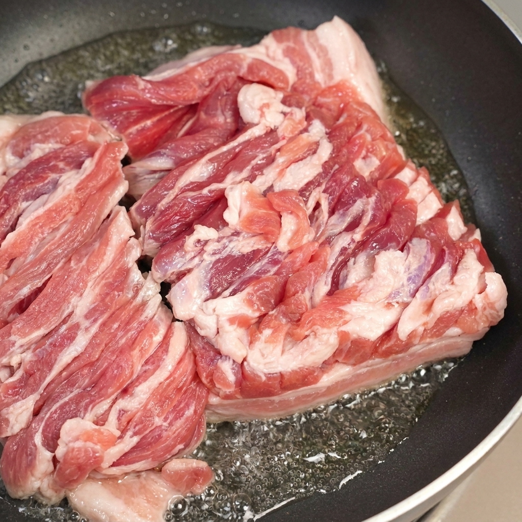 Eine Pfanne mit brutzelndem Schweinebauch und einem Löffel zum Abschöpfen des überschüssigen Fettes.