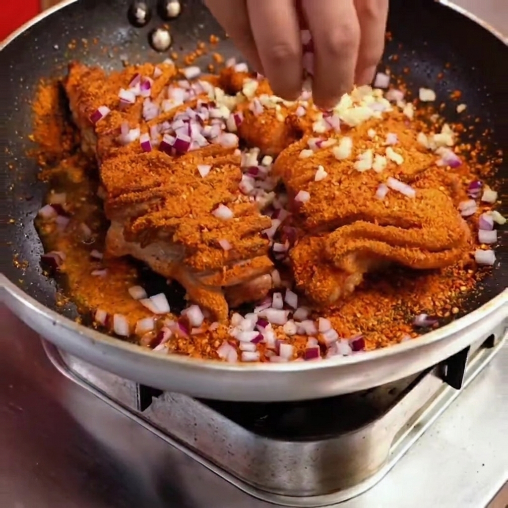 Nahaufnahme von gewürfelten roten Zwiebeln und grünen Paprikas, die über das gewürzte Schweinefleisch in die Pfanne gegeben werden.