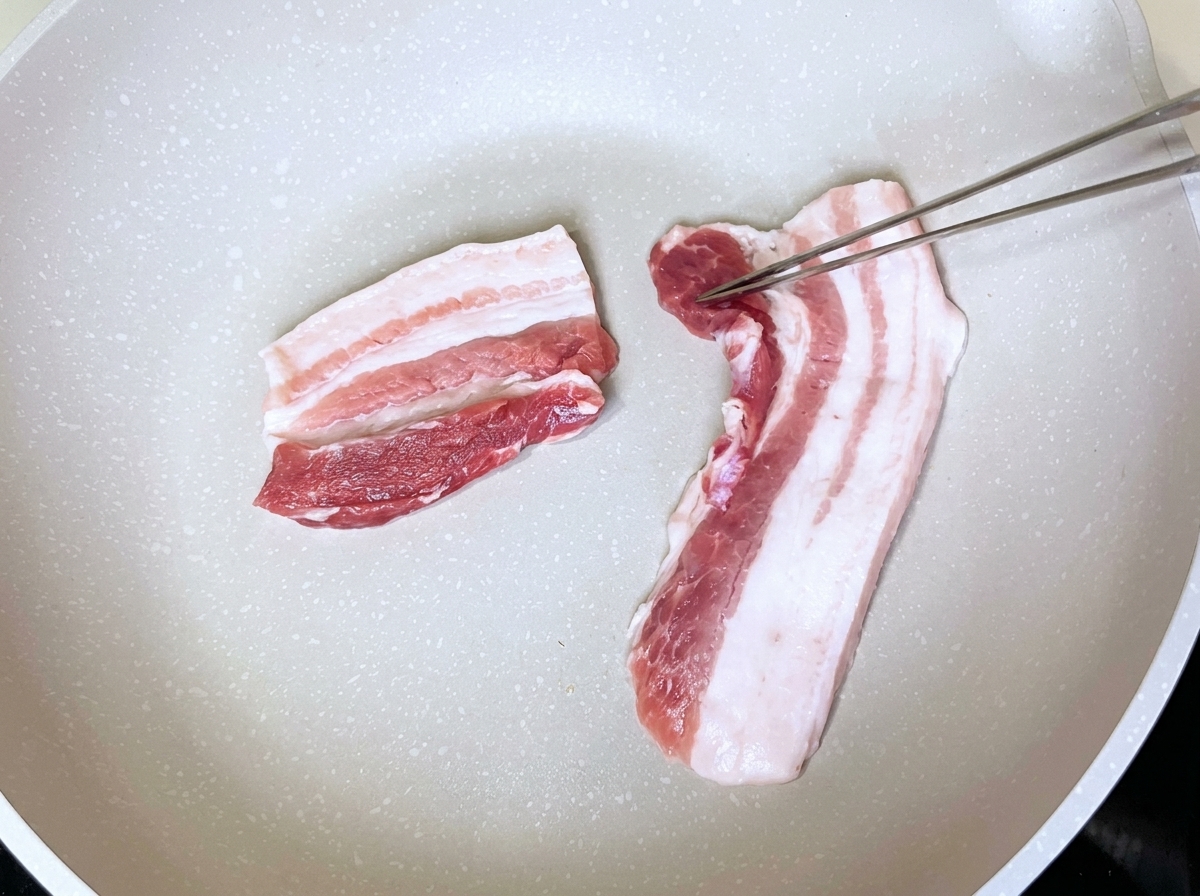 Ordentlich in einer trockenen weißen Pfanne ausgelegte Schweinebauchscheiben, die mit einer Küchenzange gewendet werden.