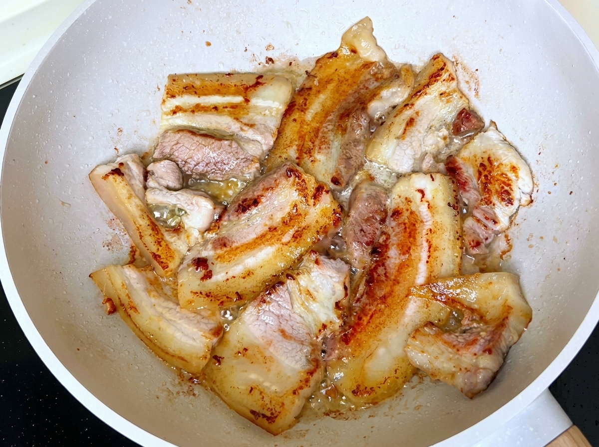 In einer Pfanne mit ausgetretenem Fett bruhende und bräunende Schweinebauchscheiben.