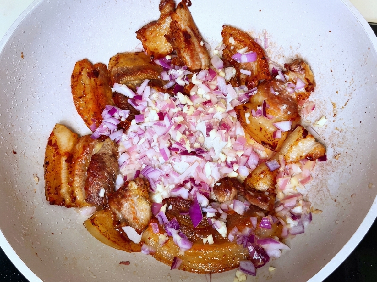 Gewürfelte rote Zwiebeln und gehackter Knoblauch über gebräunten Fleischstückchen in einer weißen Pfanne.