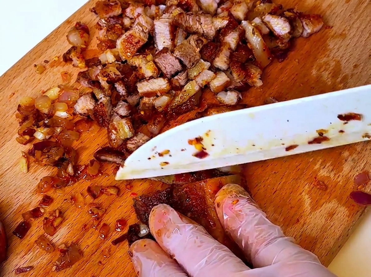 Ein Messer schneidet zubereitetes Barbecue-Fleisch und Zwiebeln auf einem Holzschneidebrett; Hände tragen Schutzhandschuhe.
