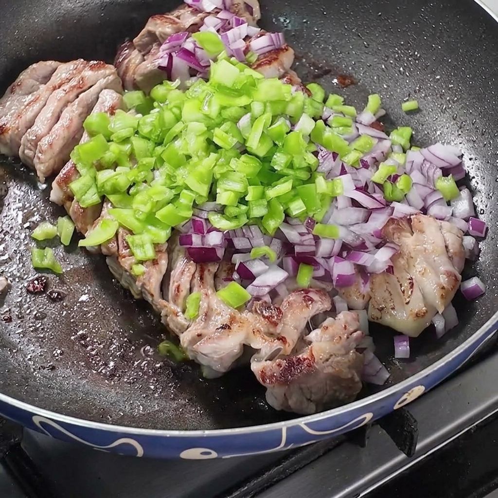 Anbratender Schweinebauch in der Pfanne mit Zwiebeln, Paprika und Gewürzen.