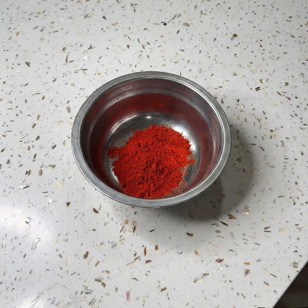 Eine kleine Metalschuessel mit einem Huegel roten Chilipulvers und weissem Zucker auf einer Granitarbeitsplatte.