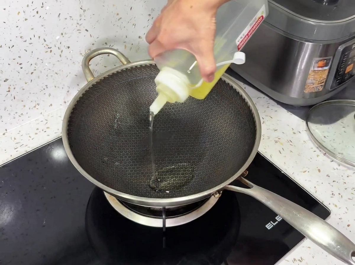 Speiseoel wird aus einer Quetschflasche in einen strukturierten Metallwok auf einem schwarzen Induktionskochfeld gegossen.