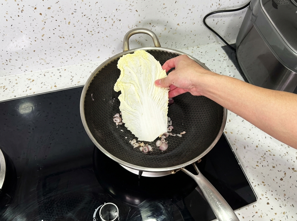 Eine Hand legt ein grossen Blatt Chinakohl in einen Wok mit gekochten Zwiebeln.