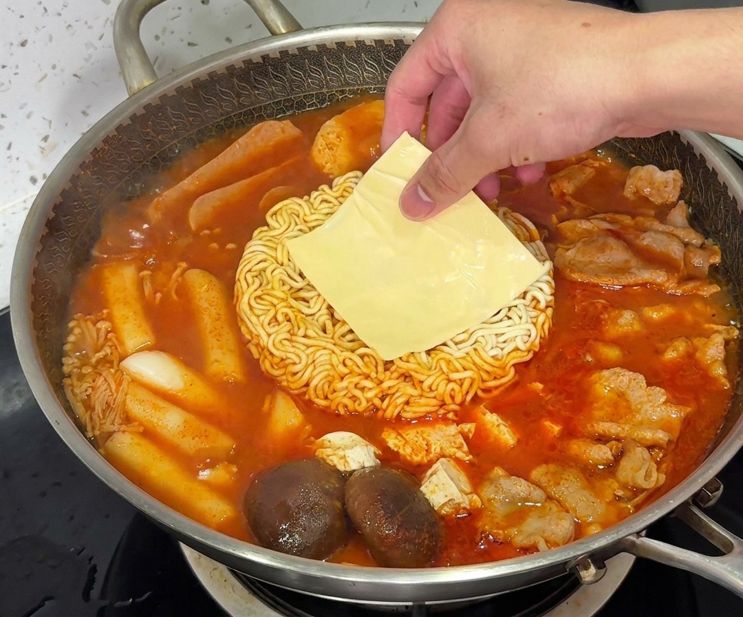 Eine Scheibe gelber Kase schmilzt ueber den Ramennudeln in einem dampfenden Topf Budae Jjigae.