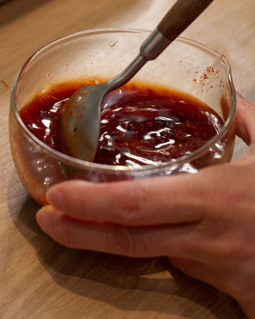 Ein Löffel rührt eine rötlich-braune Hot-Pot-Saucenmischung in einer Glasschüssel um.